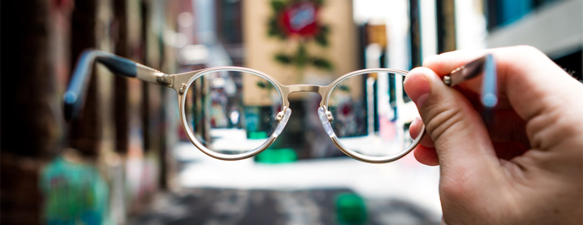 Hand holding eyeglasses in front of a blurred street scene, bringing the background into focus through the lenses.