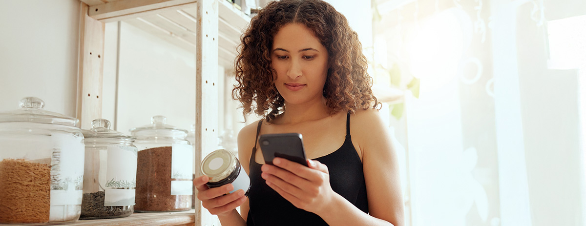 A woman stands in a bright store, holding a jar and reading its label while referencing her smartphone.