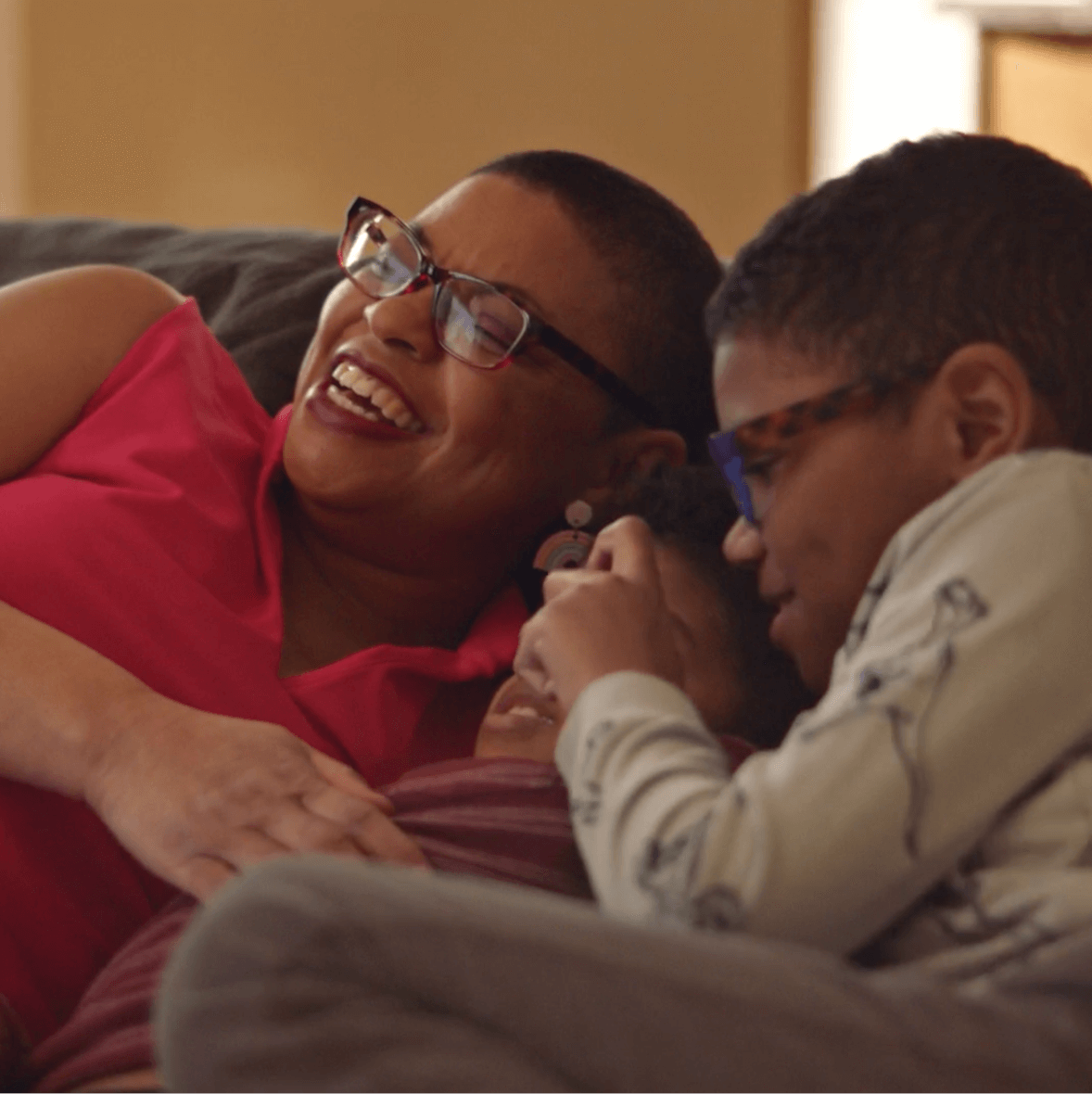 Woman with glasses and a pink shirt laughs on the couch with her 2 young children.