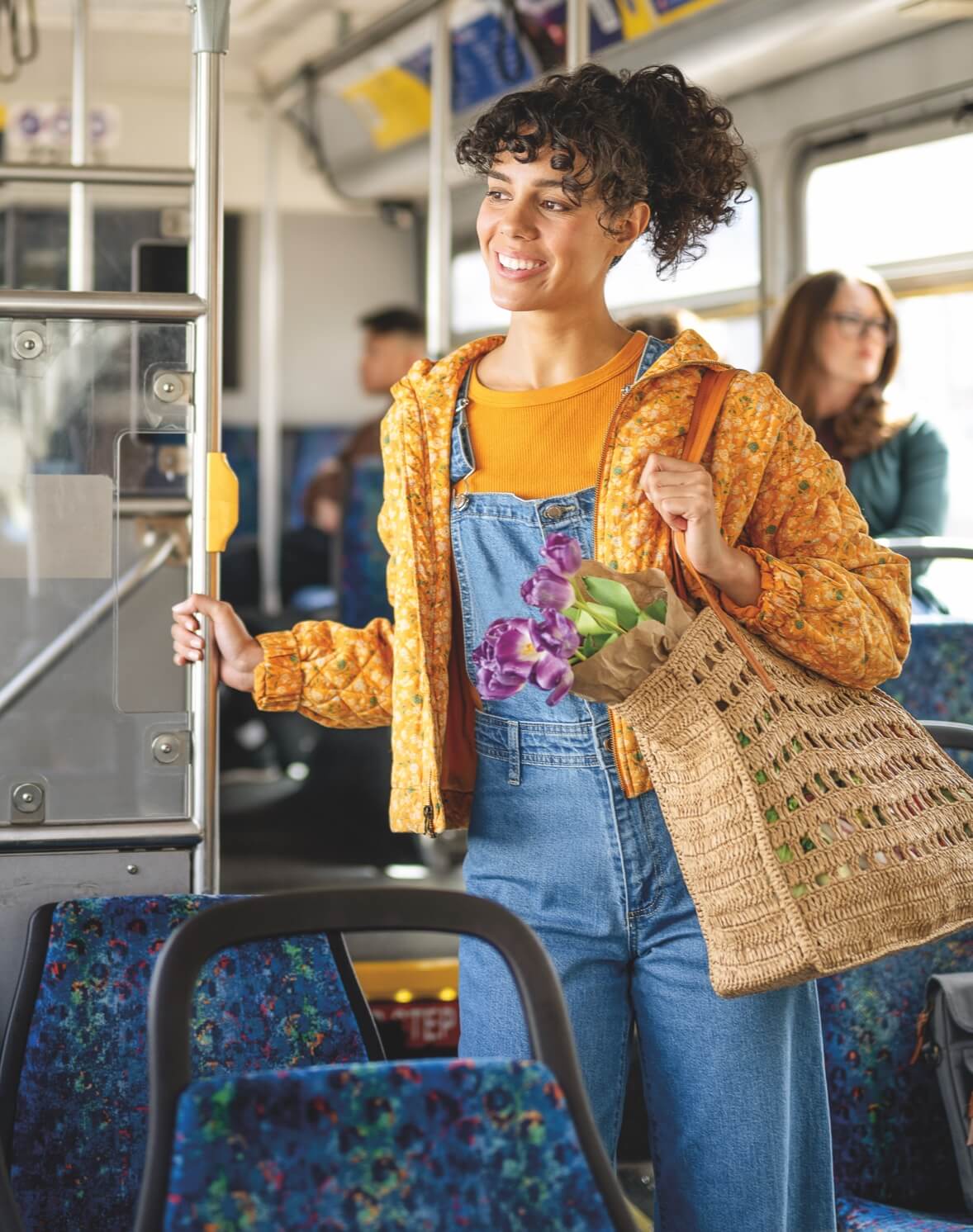 Young woman with curly brown hair, standing on a bus holding a bag with produce and flowers in it. She is smiling and wearing overalls with a floral yellow jacket.