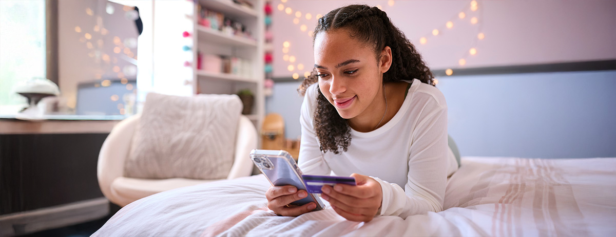 Teen lying on a bed holds a smartphone and debit card in a cozy bedroom with string lights, suggesting mobile banking or online shopping.