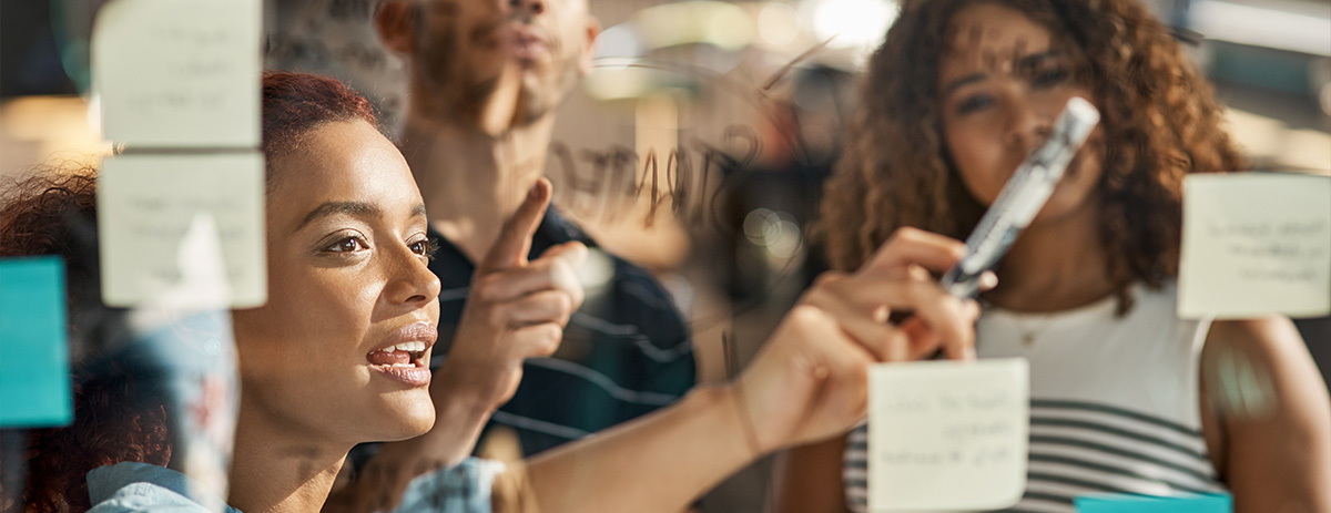 Three diverse professionals brainstorming, writing on a clear glass board with sticky notes, collaborating on ideas