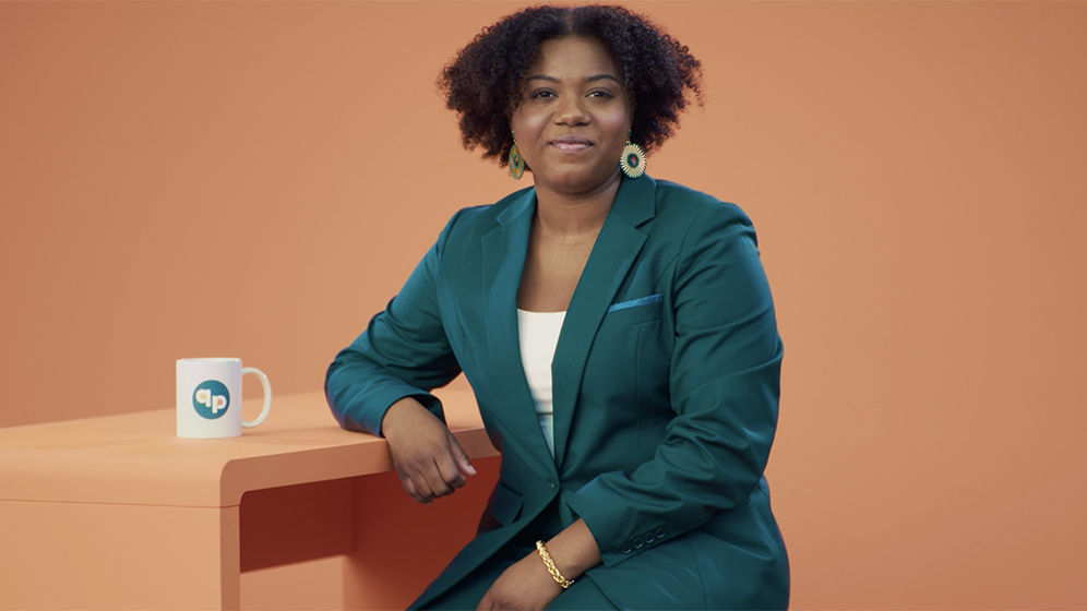 CCF_QuitPartner_VideoThumbnail A woman with short, dark curly hair sits at an orange desk against an orange background. She wears a dark teal suit and is looking straight ahead.
