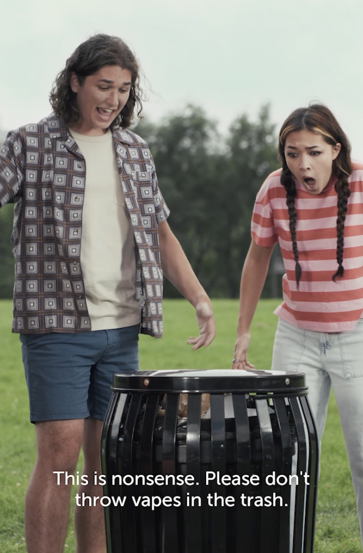 A teen boy and girl are overly expressive as the boy throws a vape into a trashcan in a city park. Caption on the image says, "This is nonsense. Please don't throw vapes in the trash."