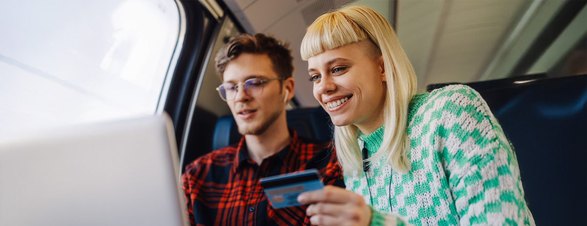 Two people on a train smiling while shopping online on a laptop, holding a credit card.