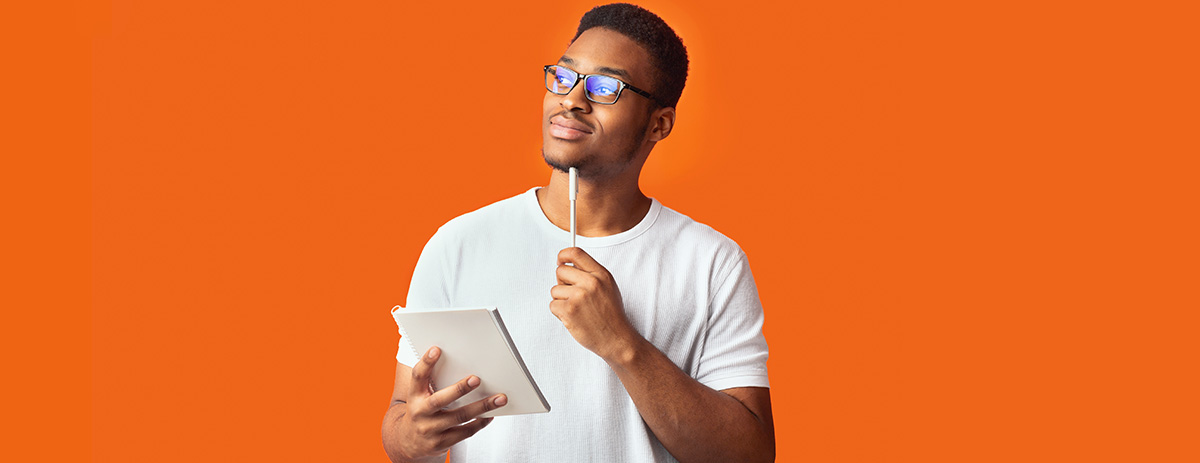Thoughtful man wearing glasses holds a notebook and pen, looking upward against a bright orange background