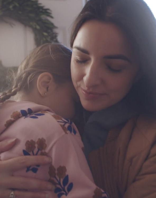 A woman gently hugs a young child wearing a pink hoodie at a home's front entryway, conveying comfort, care, and reassurance.
