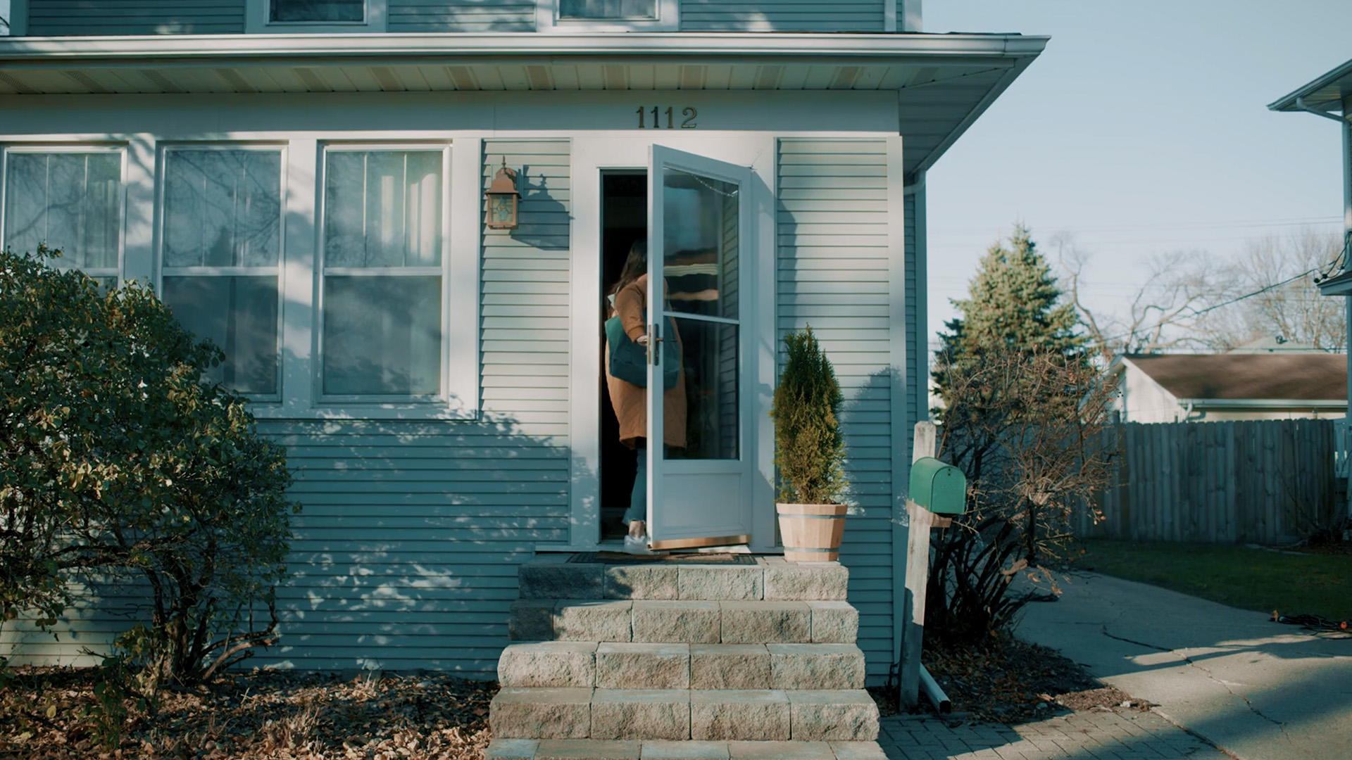 Altru_Home_Video-30 Exterior of a residential home with the front door open as a woman enters, carrying a shoulder bag up the front steps.