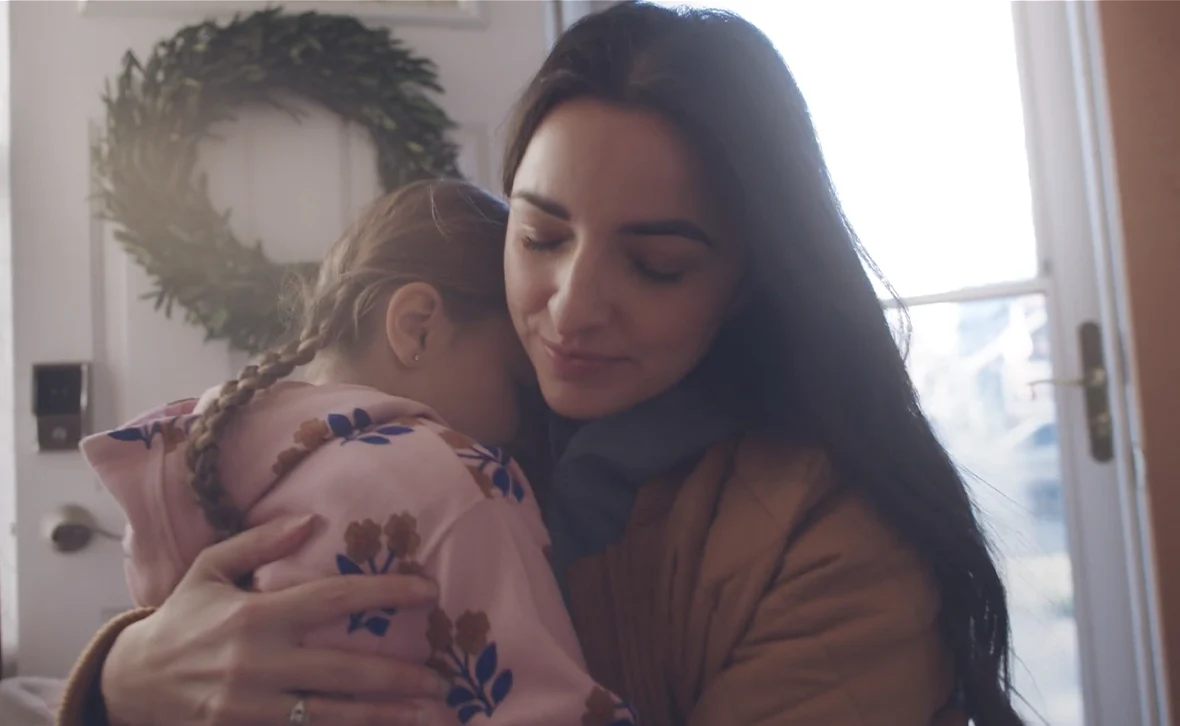 A woman gently hugs a young child wearing a pink hoodie at a home's front entryway, conveying comfort, care, and reassurance.