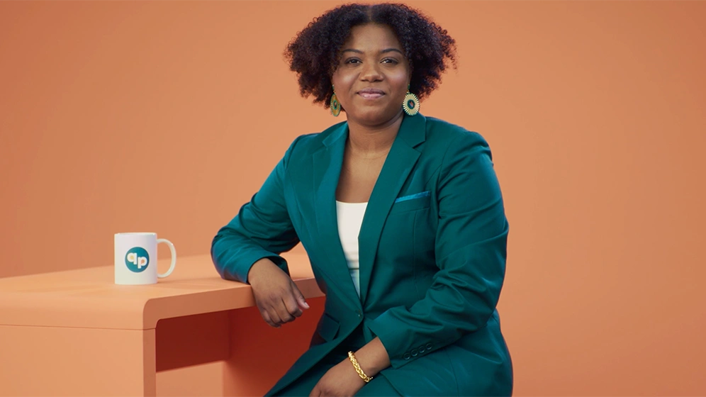 A woman with short, dark curly hair sits at an orange desk against an orange background. She wears a dark teal suit and is looking straight ahead.