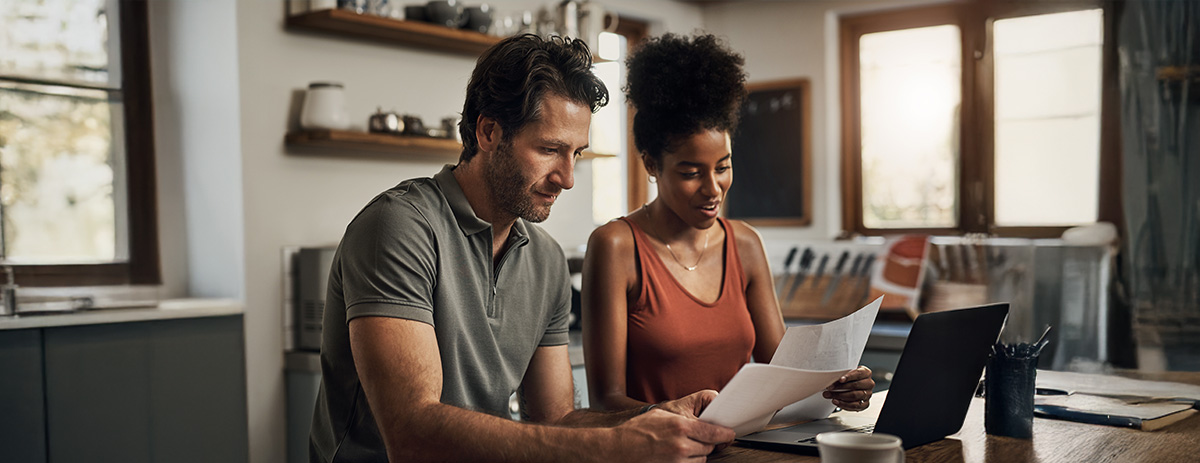 A couple reviews financial documents together on a laptop at their kitchen table at home.